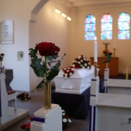Kirkepynt med rød rose i vase og lys ved kiste i kirken